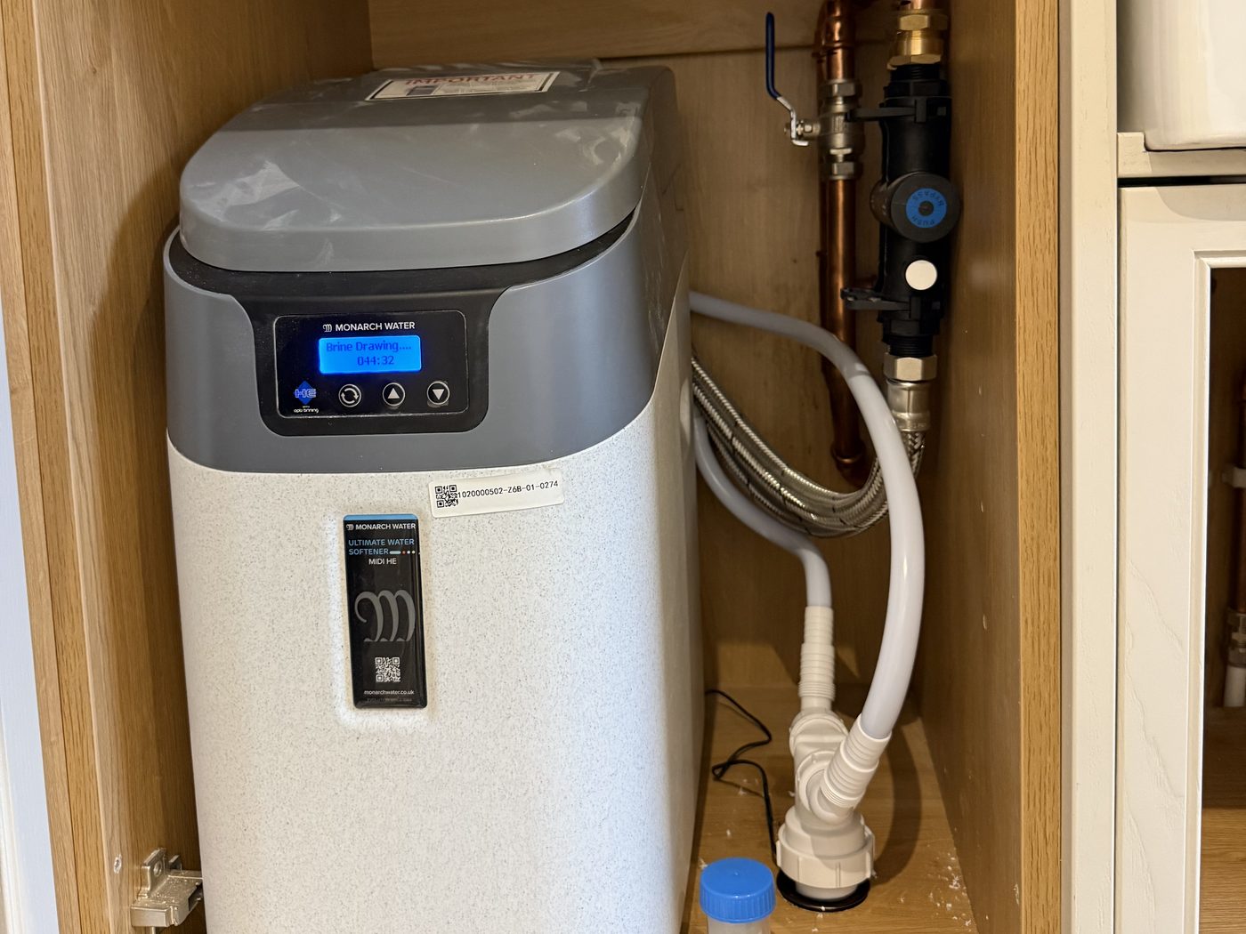 Water softener installed by Precision inside a kitchen unit, with neat pipework and bypass valve
